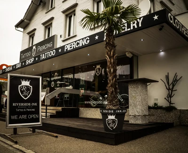 Das Haus an der Grenzstrasse 25 in St. Margrethen: die schwarze Front von Riverside Ink mit Palme vor dem Eingang. Im selben Haus ist Riverside Gastro.